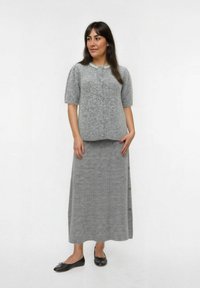 Vrouw in een grijze gebreide trui met korte mouwen en een parel-kraagdetail, gecombineerd met een bijpassende lange ribgebreide rok en zwarte ballerina's.