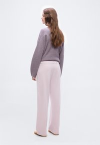 Γυναίκα με μακριά καστανά μαλλιά, που φοράει μοβ πλεκτό πουλόβερ και ανοιχτό ροζ παντελόνι με φαρδιά μπατζάκια, στέκεται μπροστά σε λευκό φόντο.