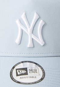 Modrá baseballová čiapka s bielou vyšitou logom "NY", nastaviteľný popruh a čierna kruhová nálepka s nápisom "NEW ERA ADJUSTABLE." Hladká textúra tkaniny.