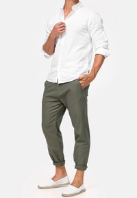 Biała lniana koszula z guzikami i podwiniętymi rękawami, w zestawieniu z oliwkowymi zwężanymi spodniami i białymi butami wsuwanymi.