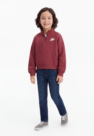 Barn med skulderlangt mørkt hår, smilende, iført en mørkerød Nike trøje, blå jeans og grå sneakers, stående foran en hvid baggrund.