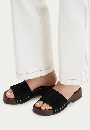 Piedi che indossano pantaloni bianchi e sandali slip-on neri con dettagli borchiati su suole marrone scuro su sfondo chiaro.