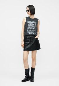 Γυναίκα που φοράει μαύρο αμάνικο μπλουζάκι με σχέδιο, μαύρη δερμάτινη φούστα με κουμπιά, μαύρες μποτάκια έως τη μέση της γάμπας και γυαλιά ηλίου, στέκεται με το ένα χέρι στην τσέπη.