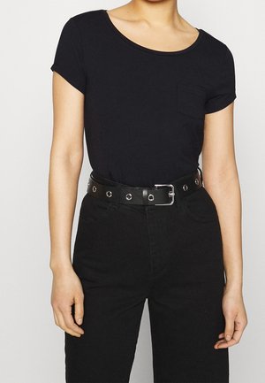 Personne portant un t-shirt noir uni à manches courtes rentré dans un pantalon noir taille haute avec une ceinture noire avec des œillets et une boucle argentés.