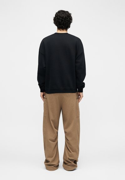 Άνδρας που στέκεται με την πλάτη στην κάμερα, φορώντας ένα μαύρο πουκάμισο με μακριά μανίκια και χαλαρά καφέ παντελόνια, μπροστά σε απλό λευκό φόντο.