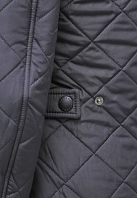 Gestepptes navy-blaues Gewebe mit einer glatten Textur. Verfügt über einen schwarzen Druckknopf und ein Akzentetikett, das auf einem rechteckigen Patch gestickt ist.