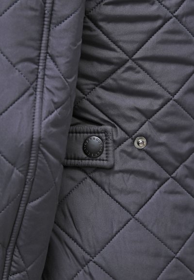 Tissu bleu marine matelassé avec une texture lisse. Présente un bouton pression noir et une étiquette décorative cousue sur un patch rectangulaire.
