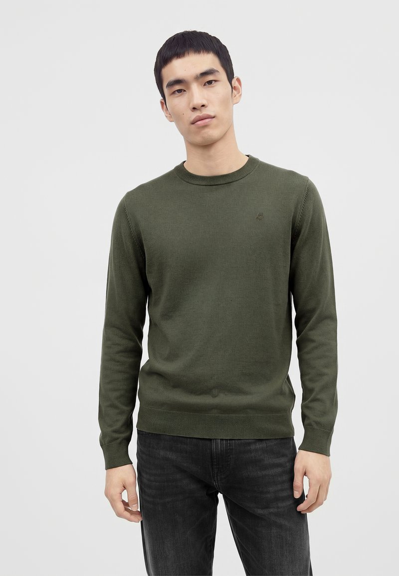 Jeune homme aux cheveux noirs courts portant un pull vert olive à manches longues et un jean gris foncé sur un fond clair uni.