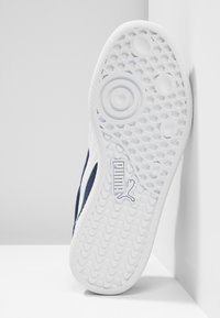 Weiße Gummisohle mit strukturiertem Muster und kreisförmigen Akzenten, mit einem Obermaterial aus navyblauem Stoff und einem sichtbaren Puma-Logo an der Seite.