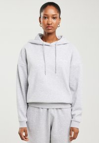 Kobieta ubrana w jasno szarą bluzę z kapturem i dopasowane spodnie dresowe, stoi na tle jednolitego jasnego tła z neutralnym wyrazem twarzy.