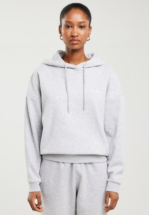 Femeie purtând un hanorac gri deschis și pantaloni de trening asortați, stând pe un fundal simplu și deschis, cu o expresie neutră.