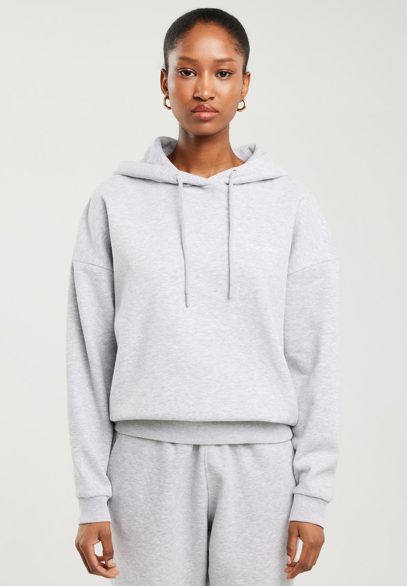 Kobieta ubrana w jasno szarą bluzę z kapturem i dopasowane spodnie dresowe, stoi na tle jednolitego jasnego tła z neutralnym wyrazem twarzy.