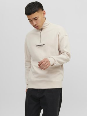 Felpa a mezzo zip color beige chiaro realizzata in tessuto morbido, con un piccolo logo nero "Originals Studio" sul petto e polsini a coste.