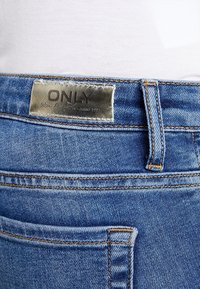 Primer plano de unos vaqueros de mezclilla azul con una etiqueta metálica de la marca "ONLY" en la cintura y detalles de costura visibles.