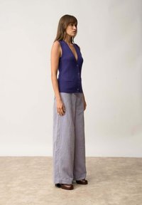 Fialová bezrukávová pletená vesta s hlubokým výstřihem do V a předními knoflíky, v kombinaci s širokými pruhovanými levandulovými kalhotami a tmavě hnědými platformovými botami.