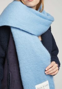 Une écharpe en laine bleu clair présente une texture douce, un design large et une étiquette avec "Andy & Lucy". Elle est portée par-dessus une veste bleu marine foncé.