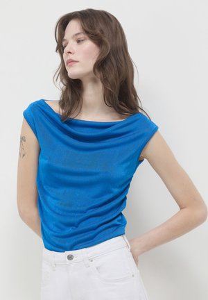 Giovane donna con lunghi capelli castani indossa un top senza maniche blu brillante e pantaloni bianchi, in piedi su uno sfondo bianco semplice.