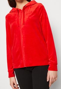 Roter Samt-Zip-up-Hoodie mit Kapuze, ausgestattet mit einer glatten Textur, Frontreißverschluss und elastischen Bündchen. Schwarze Hose ist darunter sichtbar.