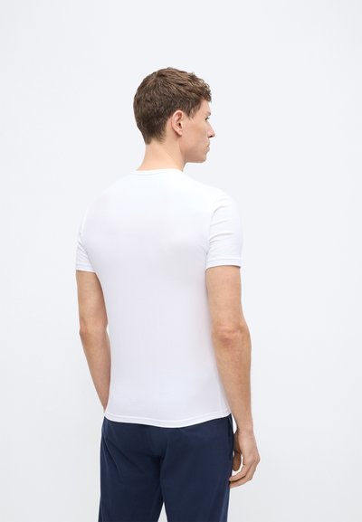 Άνδρας με λευκό κοντομάνικο μπλουζάκι και σκούρο μπλε παντελόνι, όρθιος με την πλάτη και το δεξί προφίλ του ορατά, αντιμετωπίζοντας ένα ανοιχτό γκρι φόντο.