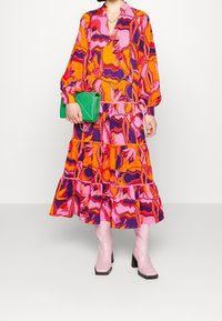 Kleurvolle maxi-jurk met een levendig bloemenpatroon in oranje, roze en paars, met ruches en lange mouwen. Groene clutch en roze laarzen.