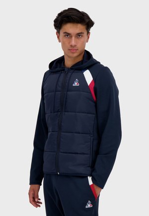 Navy padded vest with a hood, featuring a zip front, multi-color accents on the shoulders, and logo detail on the chest.