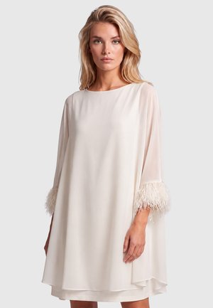Blonde woman wearing a loose cream dress with sheer three-quarter sleeves and feathered cuffs, standing against a plain background.