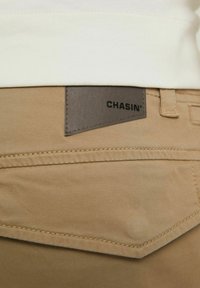 Khaki bavlněné kalhoty s béžovou koženou etiketou s nápisem "CHASIN'" a viditelnými švy. Tkanina je hladká a má volný střih.