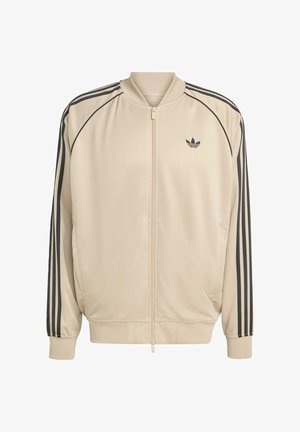 Bežová bunda na zips s čiernymi akcentmi, obsahujúca dva bočné vrecká, rebrové manžety a tri čierne pruhy pozdĺž rukávov.