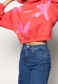 Sudadera corta color coral con un patrón abstracto en rosa, combinada con jeans de mezclilla azul de tiro alto. El material parece suave y tiene un ajuste casual.