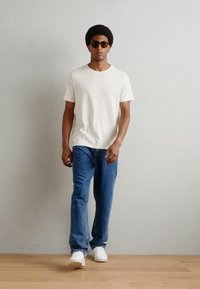 Cămașă albă cu mânecă scurtă și textură, blugi denim albastri și adidași albi. Model purtând ochelari de soare închiși la culoare, stând în fața unui fundal neutru.
