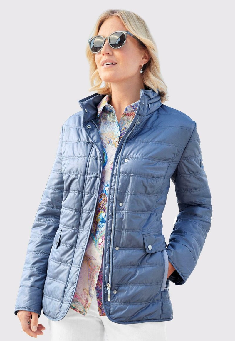 Blau gesteppte Jacke mit Stehkragen, Reißverschluss und Druckknöpfen; verfügt über Seitentaschen und eine glatte Textur. Kombiniert mit einem gemusterten Hemd.