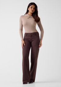 Top in maglia beige a coste con maniche lunghe e dettagli dorati; abbinato a jeans marroni a vita alta e gamba larga. Tessuto morbido, design aderente.