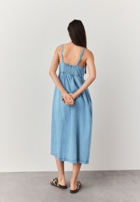 Femeie văzută de spate, purtând o rochie midi albastră din denim fără mâneci și pantofi slip-on cu imprimeu leopard, cu mâinile încheiate în spate.