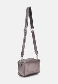 Metalická fialová kabelka z texturované kůže, obdélníkového tvaru, se zipovým uzávěrem a odnímatelným šedým popruhem s nápisem „Valentino“ černými písmeny.