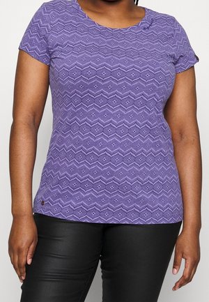 Woman wearing a purple patterned short-sleeve shirt and black pants with arms relaxed at sides against a plain background.