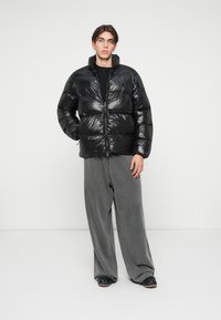 Jachetă neagră din puf cu finisaj lucios, închidere cu fermoar și guler înalt. Pantaloni gri, lejeri, completează stilul casual. Cadru minimalist.