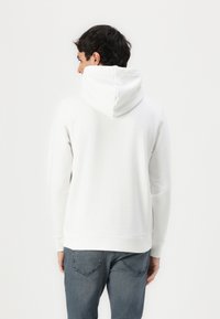 Weißer Kapuzenpullover aus weichem Material, mit Kängurutasche und gerippten Bündchen. Getragen mit dunkelblauen Jeans. Rückansicht dargestellt.