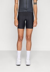 Zwarte fietsshorts met een gestructureerde tailleband, ontworpen voor comfort met een gevoerde zitting. Gepaard met witte fietsschoenen en sokken.