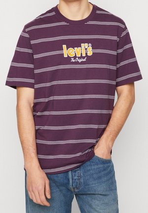 Mann trägt ein lila gestreiftes Levi's T-Shirt und blaue Jeans, mit einer Hand in der Tasche und der anderen entspannt an der Seite.