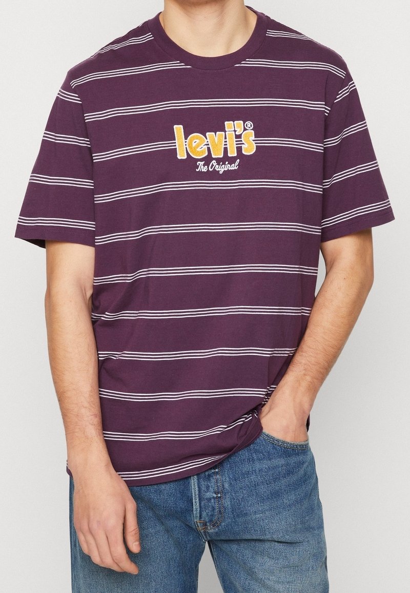 Homme portant un t-shirt Levi's rayé violet et un jean bleu, une main dans la poche et l'autre détendue sur le côté.
