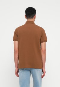 Καφέ κοντομάνικο πόλο μπλουζάκι με κλασικό γιακά, κατασκευασμέν�ο από μαλακό βαμβακερό υλικό, σε συνδυασμό με ανοιχτού μπλε τζιν παντελόνι.