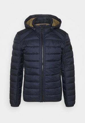Veste matelassée bleu marine avec capuche, fermeture éclair à l'avant, poches latérales zippées et doublure intérieure beige contrastante.