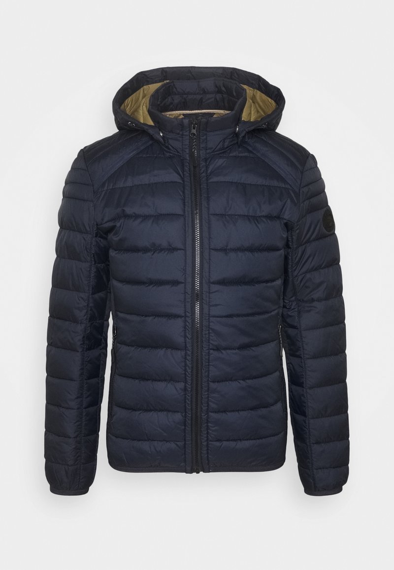 Veste matelassée bleu marine avec capuche, fermeture éclair à l'avant, poches latérales zippées et doublure intérieure beige contrastante.