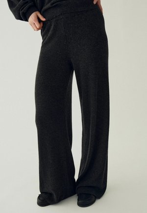 Personne portant un pantalon large ample gris foncé et des chaussures noires, une main reposant sur la hanche, sur un fond uni.