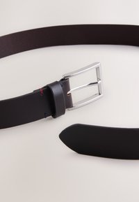 Cinturón de cuero negro con hebilla metálica plateada. Presenta un acabado liso y costuras contrastantes en rojo y azul cerca del lazo del cinturón.