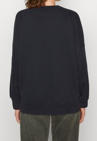 Persona che indossa un maglione nero a maniche lunghe semplice e pantaloni verde oliva, mostrata di spalle su uno sfondo neutro.