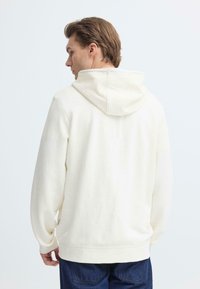 Sweat à capuche blanc en tissu doux avec une coupe décontractée, doté d'une poche kangourou et d'une capuche réglable avec cordon. Associé à un jean bleu.