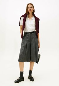 Witte polo met geborduurde details, bordeauxrode trui over de schouders, grijs gestreepte culottes, zwarte loafers en grijze sokken.