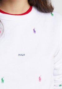 Vit sweatshirt med röd ribbad krage, prydd med små broderade polo-spelare-logotyper i lila, grönt och rosa. Texten "POLO" i marinblå.