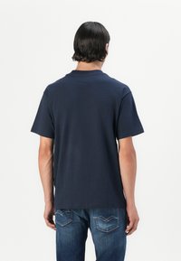 Middels blå bomull t-skjorte med korte ermer og rund hals, vist fra baksiden, kombinert med denim jeans med baklommer.